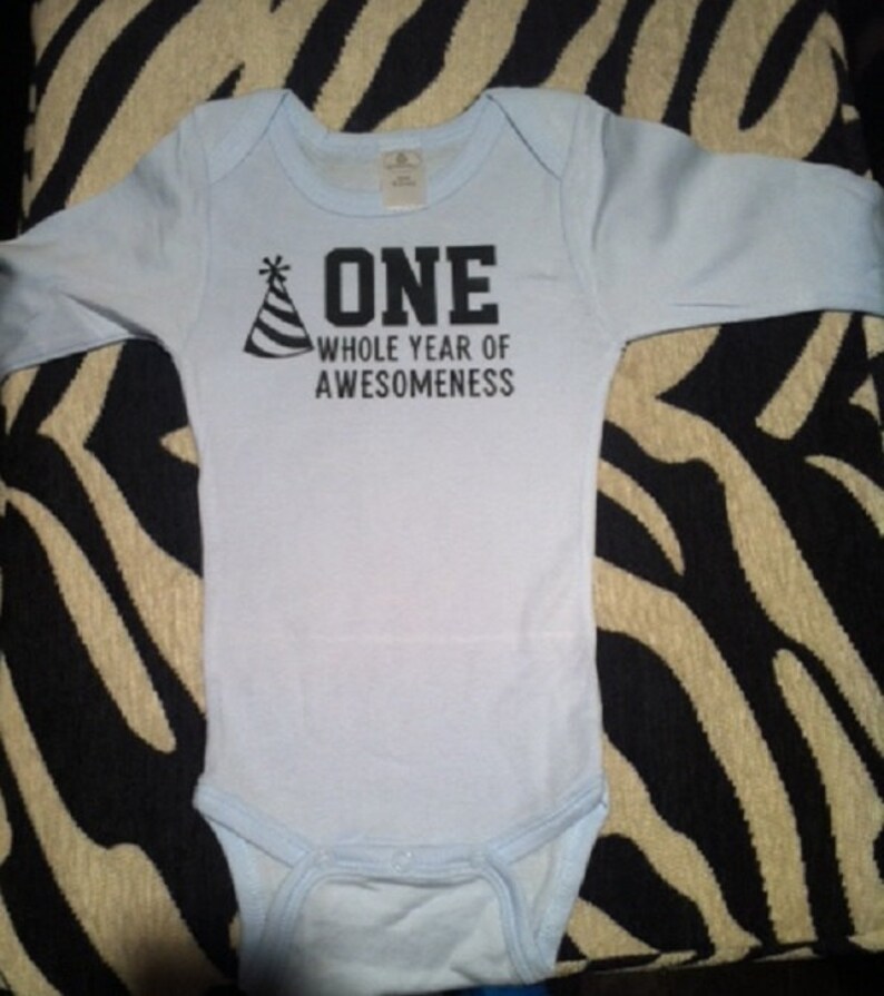 Pode incluir: Um body de beb&ecirc; azul claro com mangas compridas. O texto "ONE WHOLE YEAR OF AWESOMENESS" est&aacute; impresso em preto, acompanhado de um gr&aacute;fico de chap&eacute;u de festa. Este body &eacute; projetado para o primeiro anivers&aacute;rio de um beb&ecirc;.