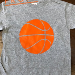 May include: A gray t-shirt with an orange basketball graphic on the front.