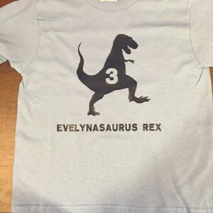 K&ouml;nnte beinhalten: Hellblaues T-Shirt mit einer schwarzen Silhouette eines Dinosauriers mit der Zahl 3 darin. Der Text "EVELYNASAURUS REX" ist unter dem Dinosaurier gedruckt.