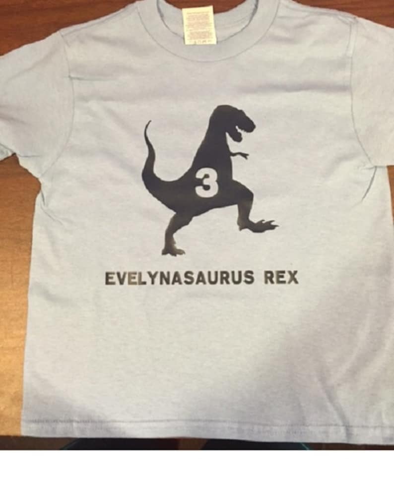 K&ouml;nnte beinhalten: Hellblaues T-Shirt mit einer schwarzen Silhouette eines Dinosauriers mit der Zahl 3 darin. Der Text "EVELYNASAURUS REX" ist unter dem Dinosaurier gedruckt.