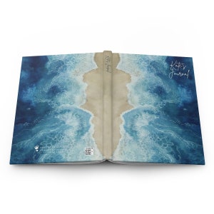 May include: A blue and brown journal with a watercolor design of ocean waves crashing on a sandy beach. The front cover has the text "Kate's Journal" in white cursive writing. The back cover has a small logo and a QR code.