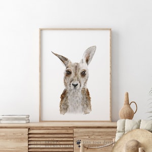 May include: A watercolor painting of a kangaroo with brown fur and a white chest. The kangaroo is looking directly at the viewer with a neutral expression. The painting is framed in a light wood frame.