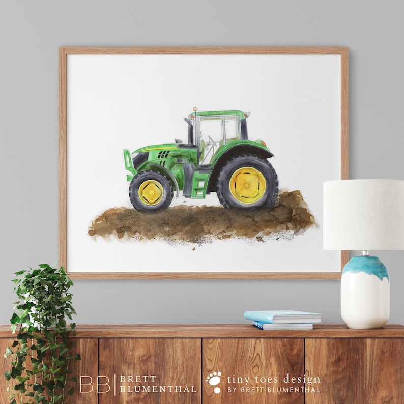 May include: Watercolor painting of a green tractor with yellow wheels, set against a white background. The tractor is depicted on a patch of brown soil, framed in a light brown wooden frame. The artwork is a print.