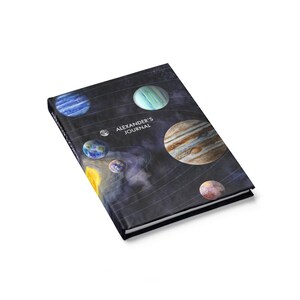 May include: A black journal with a galaxy design featuring planets in shades of blue, yellow, brown, and red. The text "ALEXANDER'S JOURNAL" is printed in white on the front cover.