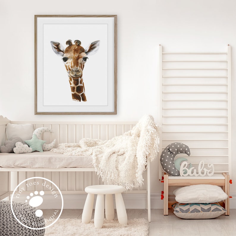 K&ouml;nnte beinhalten: Ein gerahmtes Aquarellbild eines Giraffenkopfes vor wei&szlig;em Hintergrund. Der Rahmen ist aus hellem braunem Holz. Das Bild h&auml;ngt an einer wei&szlig;en Wand &uuml;ber einem wei&szlig;en Kinderbett mit einer wei&szlig;en Decke und einem wei&szlig;en Hocker. Das Kinderbett hat ein wei&szlig;es und graues sternf&ouml;rmiges Kissen und ein hellblaues Halbmondkissen. Der Hocker steht vor einem wei&szlig;en Teppich. Die Wand hinter dem Kinderbett hat ein wei&szlig;es Leiterstellregal mit einem wei&szlig;en Holzschild, auf dem "Baby" steht.