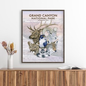 The image is a framed poster of various animals, including a deer, a bird, a wolf, and a cat, all set against a pale pink background. The poster is hanging on a wall, and there is a vase of flowers and a book on a wooden cabinet below it.