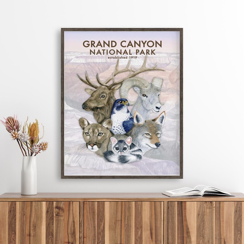 The image is a framed poster of various animals, including a deer, a bird, a wolf, and a cat, all set against a pale pink background. The poster is hanging on a wall, and there is a vase of flowers and a book on a wooden cabinet below it.
