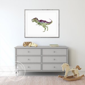 Pode incluir: Uma pintura em aquarela de um dinossauro verde e roxo em uma moldura branca pendurada em uma parede branca acima de uma c&ocirc;moda cinza com seis gavetas. H&aacute; dois carrinhos de brinquedo de madeira no topo da c&ocirc;moda e um cavalinho de balan&ccedil;o de madeira na frente da c&ocirc;moda.