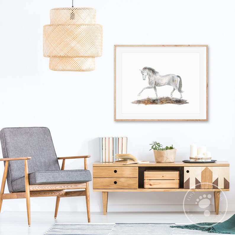 May include: A watercolor painting of a white horse running on a brown background. The painting is framed in a light wood frame and is hanging on a white wall. A gray armchair, a wooden side table, and a potted plant are also in the image.