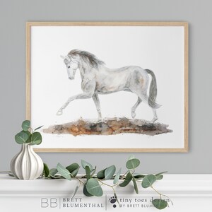 May include: A watercolor painting of a white horse running on a brown dirt path. The horse has a long, flowing mane and tail. The painting is framed in a light wood frame.