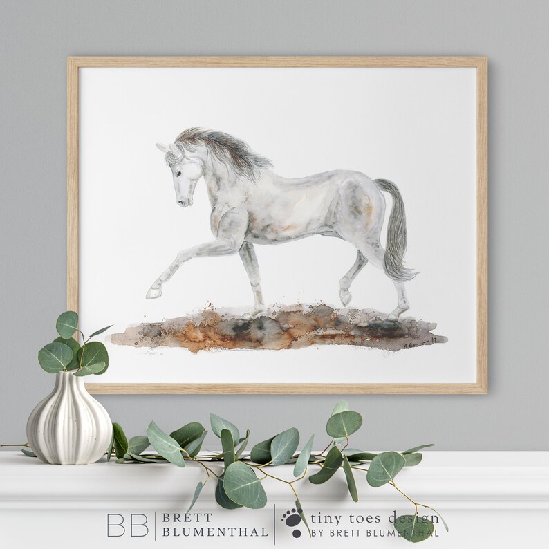 May include: A watercolor painting of a white horse running on a brown dirt path. The horse has a long, flowing mane and tail. The painting is framed in a light wood frame.