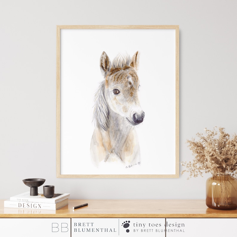 May include: A watercolor painting of a brown and white foal with a white mane and tail. The foal is looking to the right of the image. The painting is framed in a light wood frame.
