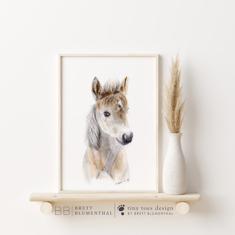 May include: A watercolor painting of a light brown and white foal with a white mane and tail. The foal is looking to the right of the image. The painting is framed in a simple white frame.