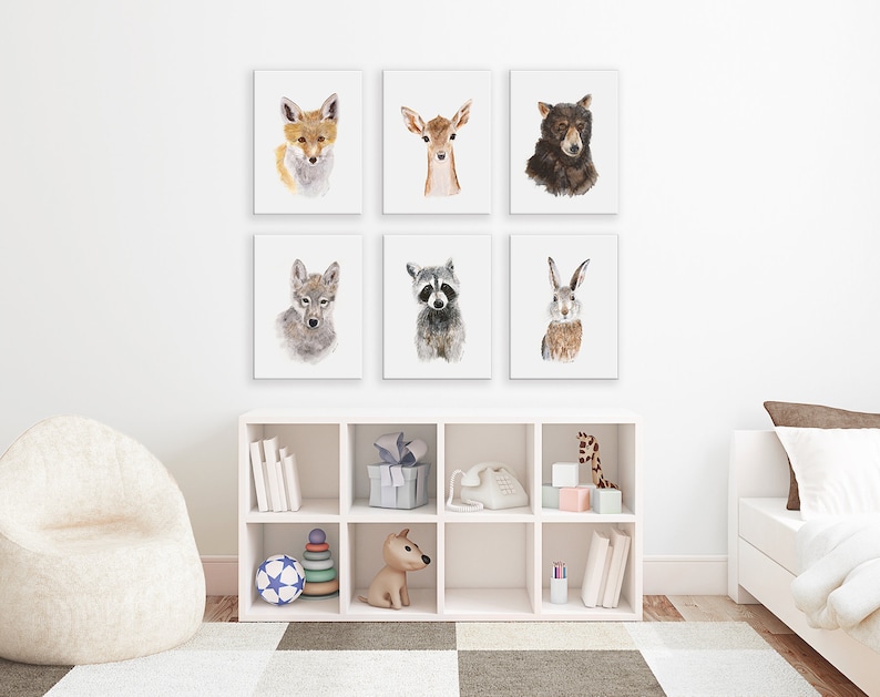 Pu&ograve; includere: Un set di sei stampe di animali ad acquerello che raffigurano una volpe, un cervo, un orso, un lupo, un procione e un coniglio. Le stampe sono esposte su una parete bianca in una cameretta o in una nursery.