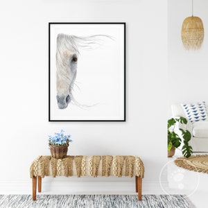 May include: A watercolour painting of a white horse's head with a black frame. The horse's eye is brown and the mane is flowing in the wind. The painting is hanging on a white wall above a light brown bench with fringe.