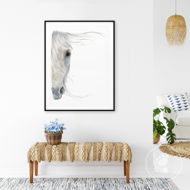 May include: A watercolour painting of a white horse's head with a black frame. The horse's eye is brown and the mane is flowing in the wind. The painting is hanging on a white wall above a light brown bench with fringe.