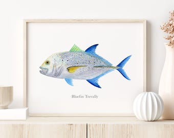 May include: Watercolor print of a Bluefin Trevally fish. The fish has a light blue body with darker blue fins and a yellow stripe. The artwork is framed in a light wood frame and is displayed on a white background.