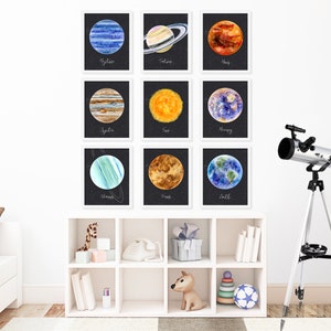 以下が含まれることがあります： 白い壁に、白い額縁に入った太陽系の惑星の水彩画が9枚飾られています。惑星は、海王星、土星、火星、木星、太陽、水星、天王星、金星、地球です。絵画は3行3列に配置されています。画像の右下には、銀色の望遠鏡が三脚に載っています。