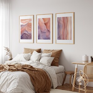 K&ouml;nnte beinhalten: Drei gerahmte abstrakte Kunstdrucke im Aquarell-Stil. Die Drucke zeigen eine Vielzahl von Farben, darunter Rosa, Lila und Orange. Die Drucke h&auml;ngen an einer wei&szlig;en Wand &uuml;ber einem Bett mit braunen Bettw&auml;sche.
