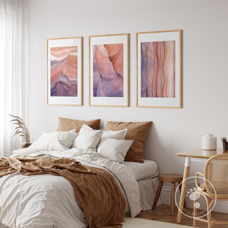K&ouml;nnte beinhalten: Drei gerahmte abstrakte Kunstdrucke im Aquarell-Stil. Die Drucke zeigen eine Vielzahl von Farben, darunter Rosa, Lila und Orange. Die Drucke h&auml;ngen an einer wei&szlig;en Wand &uuml;ber einem Bett mit braunen Bettw&auml;sche.