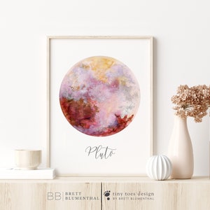 May include: A watercolor painting of Pluto, the dwarf planet, in shades of pink, red, and gold. The painting is framed and has the word "Pluto" written in cursive below the planet.