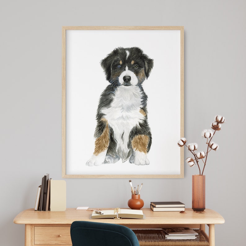 May include: A watercolor painting of a Bernese Mountain Dog puppy sitting and looking at the viewer. The puppy has black, white, and brown fur. The painting is framed in a light wood frame.