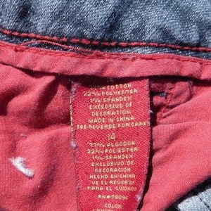 May include: A red and white clothing label with the text "COTTON 22% POLYESTER 1% SPANDEX EXCLUSIVE OF DECORATION MADE IN CHINA SEE REVERSE FOR CARE 14 77% ALGODON 22% FOLIESTER 1% SPANDEX EXCLUSIVO DE DECORACION HECHO EN CHING VEEL REVERSE PARA EL CUIDASO RM#78096 COLOR"