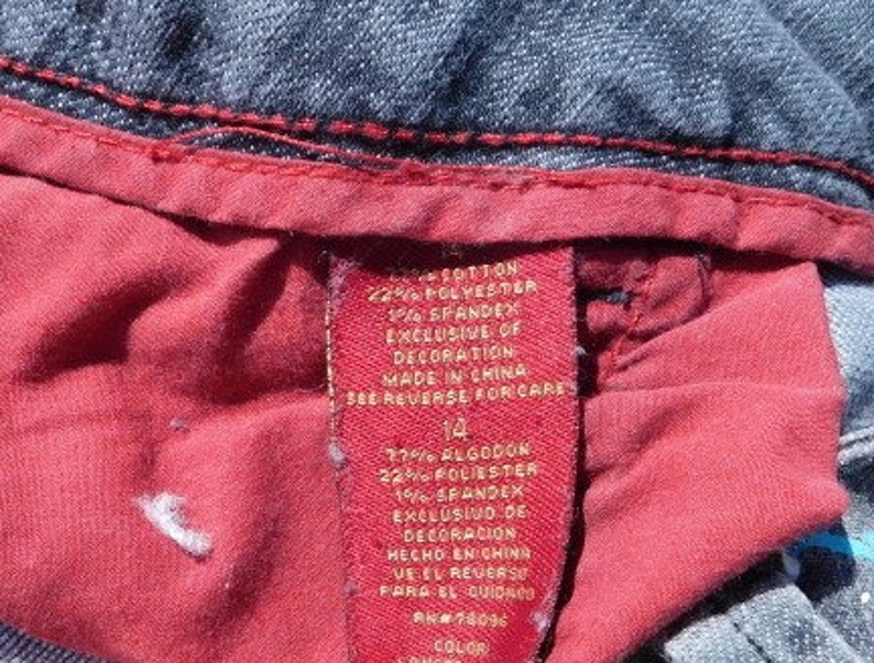 May include: A red and white clothing label with the text "COTTON 22% POLYESTER 1% SPANDEX EXCLUSIVE OF DECORATION MADE IN CHINA SEE REVERSE FOR CARE 14 77% ALGODON 22% FOLIESTER 1% SPANDEX EXCLUSIVO DE DECORACION HECHO EN CHING VEEL REVERSE PARA EL CUIDASO RM#78096 COLOR"