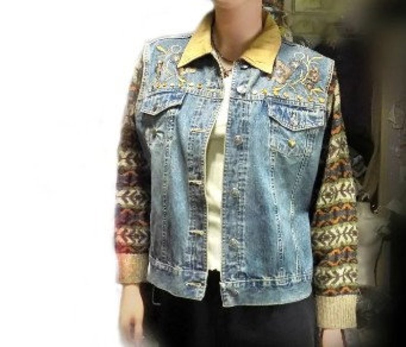 May include: A blue denim jacket with embroidered floral details on the front and a brown suede collar. The sleeves are made of a colorful knitted fabric with a geometric pattern.