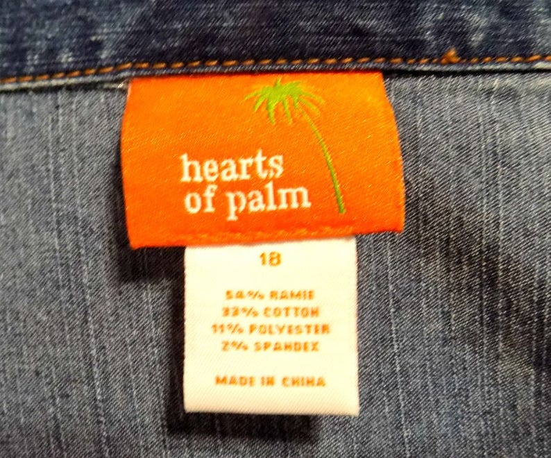 Może przedstawiać: Biała metka z czarnym tekstem, na kt&oacute;rej widnieje napis "hearts of palm" i "18" na pomarańczowej metce z grafiką palmy. Metka zawiera r&oacute;wnież informacje o składzie tkaniny: 54% Ramie, 32% Bawełna, 11% Poliester, 2% Elastan. Metka jest przymocowana do niebieskiego materiału dżinsowego.