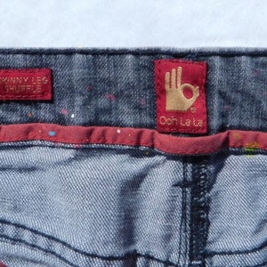 May include: Blue denim jeans with a red waistband and two red labels. The labels say "Skinny Leg Shuffle" and "Ooh La La".