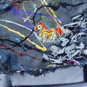 May include: A pair of blue denim jeans with colorful paint splatters and a cartoon horse patch. The jeans have distressed areas and a frayed hem.