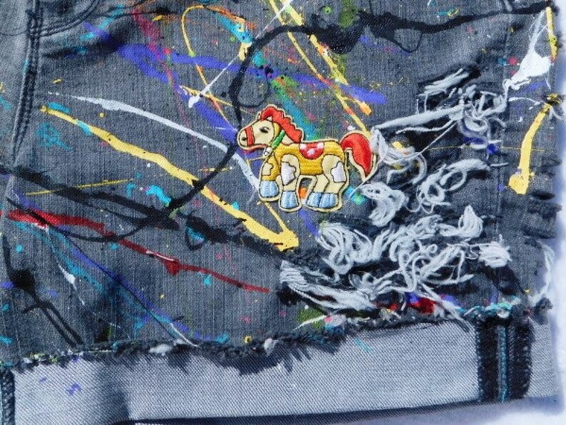 May include: A pair of blue denim jeans with colorful paint splatters and a cartoon horse patch. The jeans have distressed areas and a frayed hem.