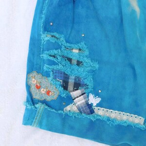 May include: A pair of blue denim shorts with distressed details, featuring a patchwork design with plaid fabric and embroidered floral accents. The shorts have a frayed hem and a white lace trim.
