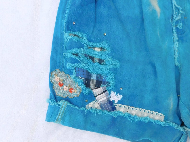 May include: A pair of blue denim shorts with distressed details, featuring a patchwork design with plaid fabric and embroidered floral accents. The shorts have a frayed hem and a white lace trim.
