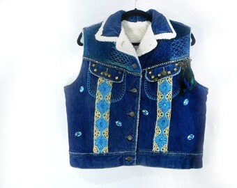 May include: A denim vest with a white sherpa lining and a blue collar. The vest is embellished with blue and gold floral embroidery, blue gemstones, and gold studs. A black hanger supports the vest.