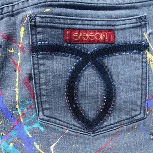 May include: Gray denim jeans with paint splatters in various colors, including yellow, blue, red, and pink. The back pocket has a black sequined design and a red label with the word "GASSION" in gold lettering.