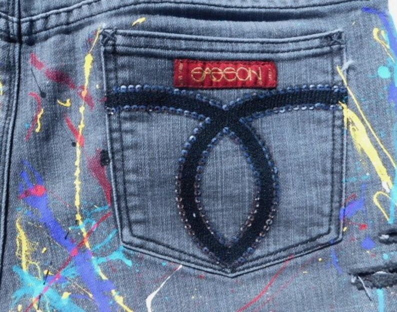 May include: Gray denim jeans with paint splatters in various colors, including yellow, blue, red, and pink. The back pocket has a black sequined design and a red label with the word "GASSION" in gold lettering.