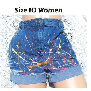 May include: Blue denim shorts with paint splatters in yellow, orange, purple, and red. The shorts have a frayed hem and a high waist.