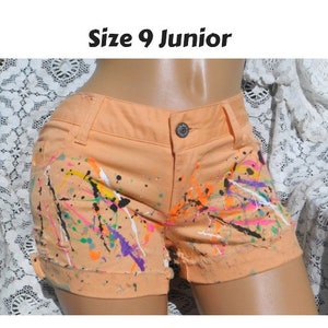 May include: Peach-colored denim shorts with a paint splatter design in various colors, including black, white, pink, green, and purple. The shorts have a size tag that reads "Size 9 Junior".