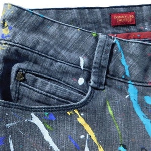 May include: Black denim jeans with a paint splatter design in various colors, including blue, yellow, white, and red. The jeans have a front pocket and a label that reads "Skinny Leg Shuffle".