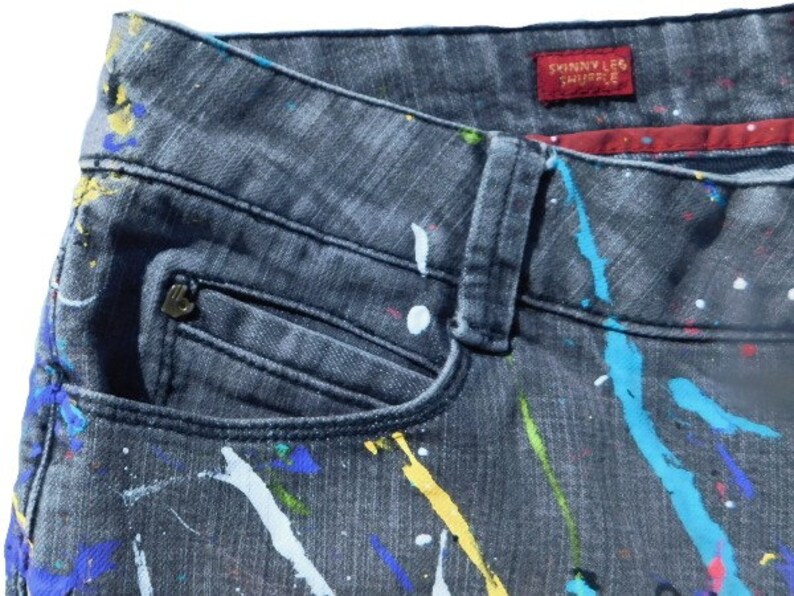 May include: Black denim jeans with a paint splatter design in various colors, including blue, yellow, white, and red. The jeans have a front pocket and a label that reads "Skinny Leg Shuffle".