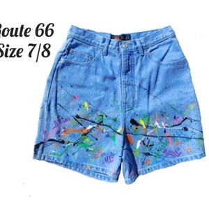 May include: Light blue denim shorts with colorful paint splatter design. The shorts have a classic five-pocket design and a button and zipper closure. The text "Route 66 Size 7/8" is visible on the upper left.