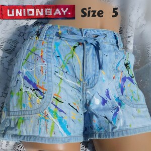 May include: Light blue denim shorts with colorful paint splatter design. Features a drawstring waist and front pockets. The shorts are size 5 and have the brand name "UNIONBAY" on a red tag.