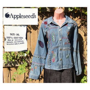 Puede incluir: Chaqueta vaquera azul con bordados florales y de hojas en rojo, verde y marrón. La chaqueta tiene detalles de tachuelas metálicas y la etiqueta "Appleseed's", talla XL. Fabricada en 100% algodón.
