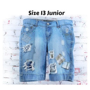 Könnte beinhalten: Hellblaue Jeansshorts mit Used-Details und dekorativen Patches. Die Shorts haben einen Knopf- und Reißverschluss, Taschen und Verzierungen. Der Text "Size 13 Junior" wird oben angezeigt.