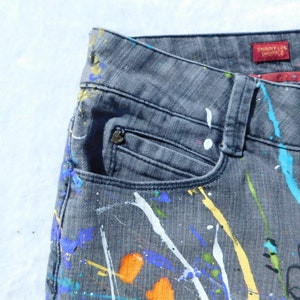 May include: Gray denim jeans with paint splatters in various colors, including white, blue, yellow, and orange. The jeans have a pocket on the left side and a red label that says "Skinny Leg Trouser".
