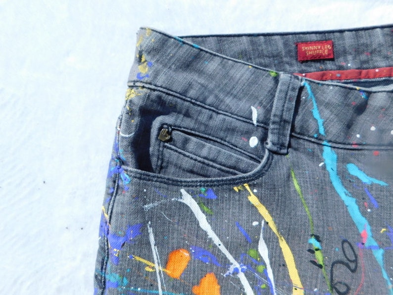 May include: Gray denim jeans with paint splatters in various colors, including white, blue, yellow, and orange. The jeans have a pocket on the left side and a red label that says "Skinny Leg Trouser".
