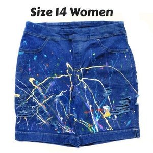 May include: Size 14 denim shorts with paint splatter design. The blue shorts feature colorful paint splatters in white, yellow, purple, and other colors. The shorts have distressed details and pockets. The text "Size 14 Women" is at the top.