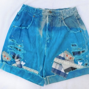 May include: Blue denim shorts with distressed details, patches, and embroidery. The shorts have a high waist and a relaxed fit.
